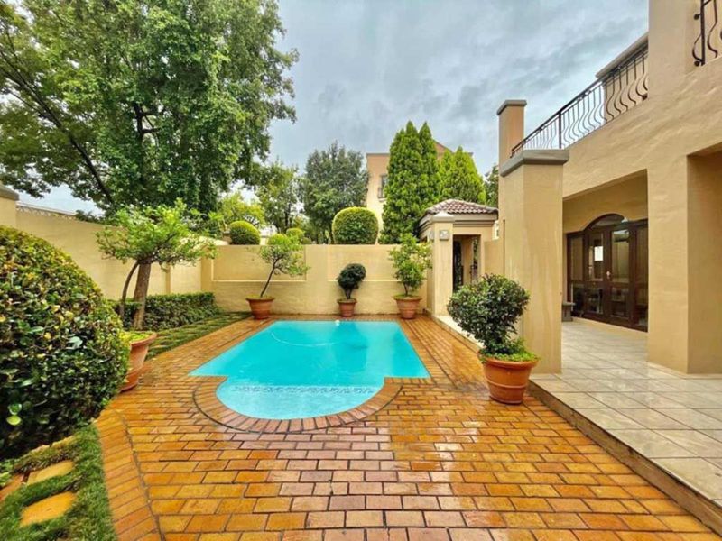 A well-maintained outdoor pool area featuring brick paving, a swimming pool, and a covered patio with a wrought iron balustrade.