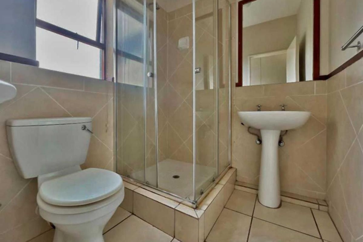 A functional bathroom featuring a pedestal basin, toilet, and a glass-enclosed shower with neutral wall and floor tiling.