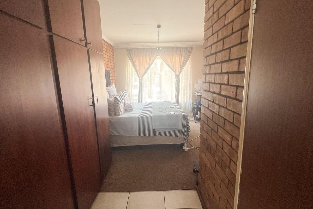 A bedroom featuring built-in wooden cupboards, an exposed brick feature wall, and a combination of tiled and carpeted flooring.