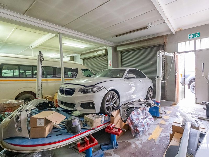 A functional workshop or garage space featuring roller shutter doors, industrial lighting, and concrete flooring, currently utilized for vehicle maintenance and repair.