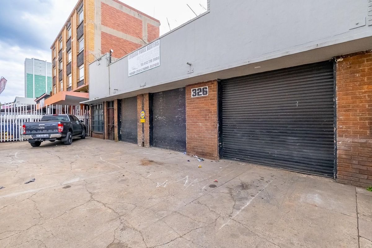 Front elevation of a commercial retail property featuring multiple roller shutter doors, brick facade, and a concrete driveway area.
