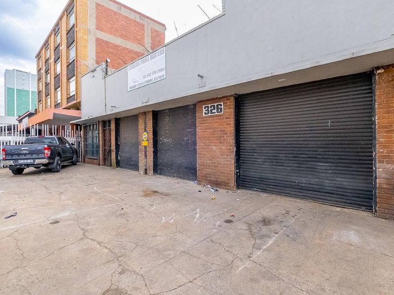 Front elevation of a commercial retail property featuring multiple roller shutter doors, brick facade, and a concrete driveway area.