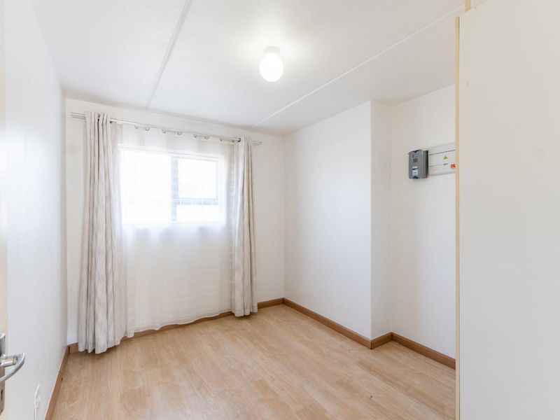 A bright, empty bedroom featuring light-toned laminate flooring, neutral walls, and a wall-mounted electrical distribution board.