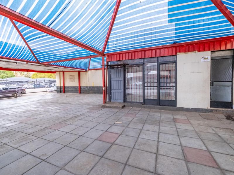Covered outdoor retail entrance area featuring a metal frame canopy with shade cloth and tiled flooring, providing a sheltered access point.
