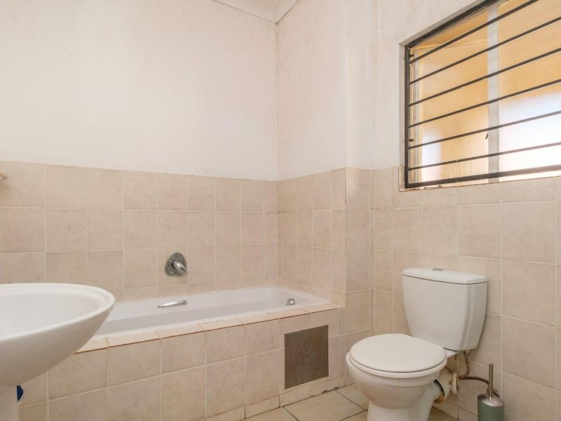 A functional bathroom featuring a bathtub, pedestal sink, and toilet with neutral wall tiling and a window with security bars.