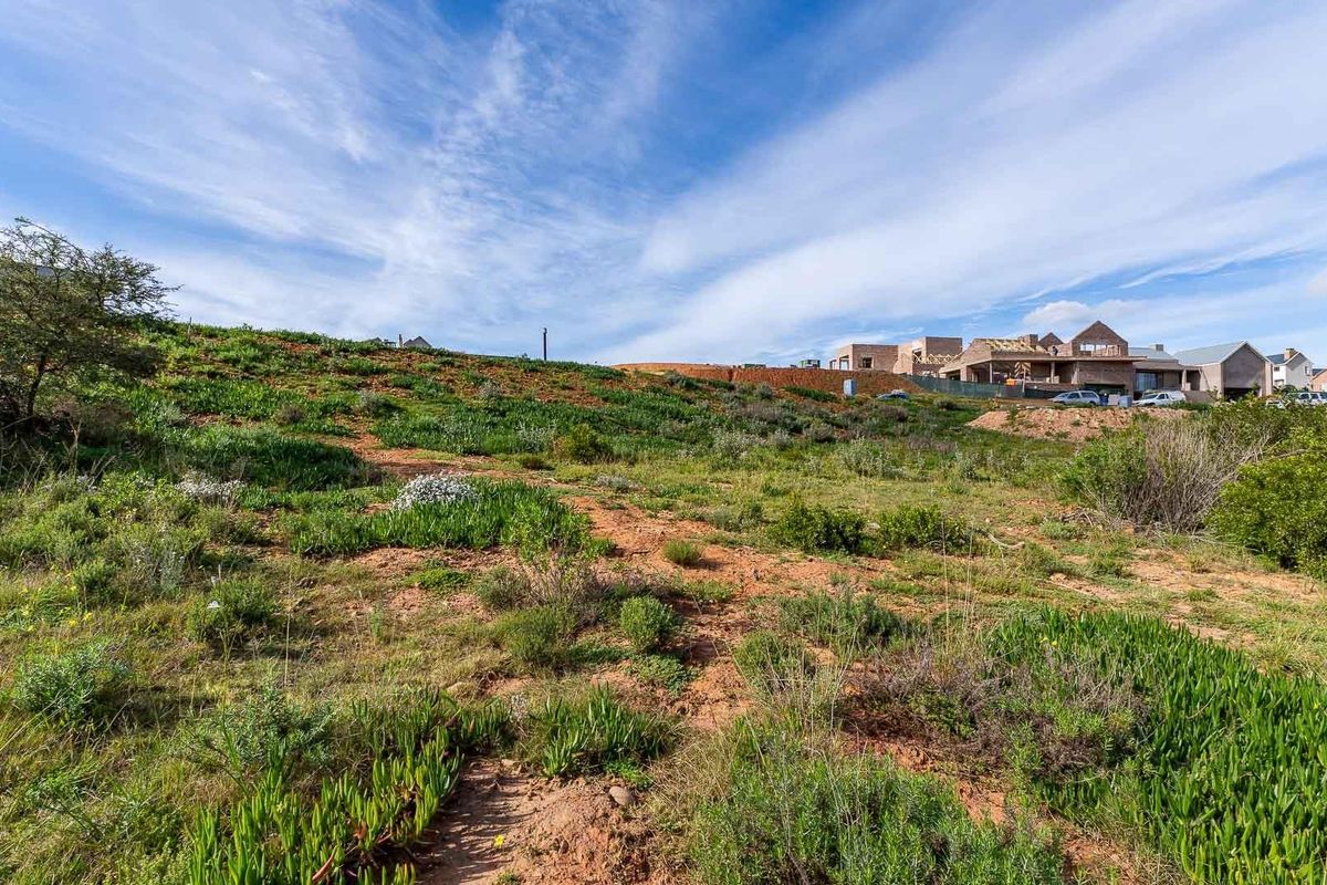 A view of a sloped, undeveloped residential plot featuring natural vegetation and surrounding neighborhood development in the background.