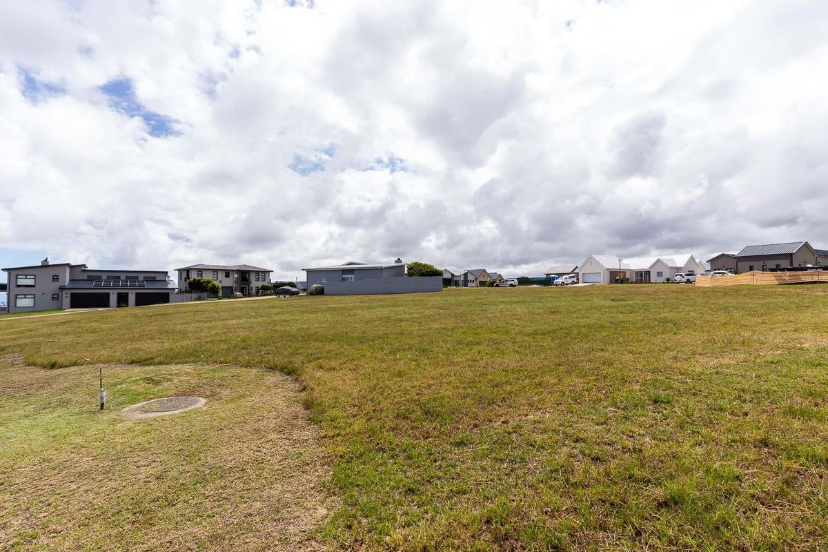 A wide-angle view of a vacant residential plot featuring open grass land with a manhole cover and utility pipe visible in the foreground.