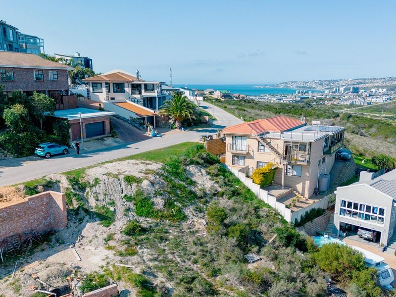 Aerial view of a vacant residential plot featuring sloped terrain and a retaining wall, situated in a developed coastal neighborhood.
