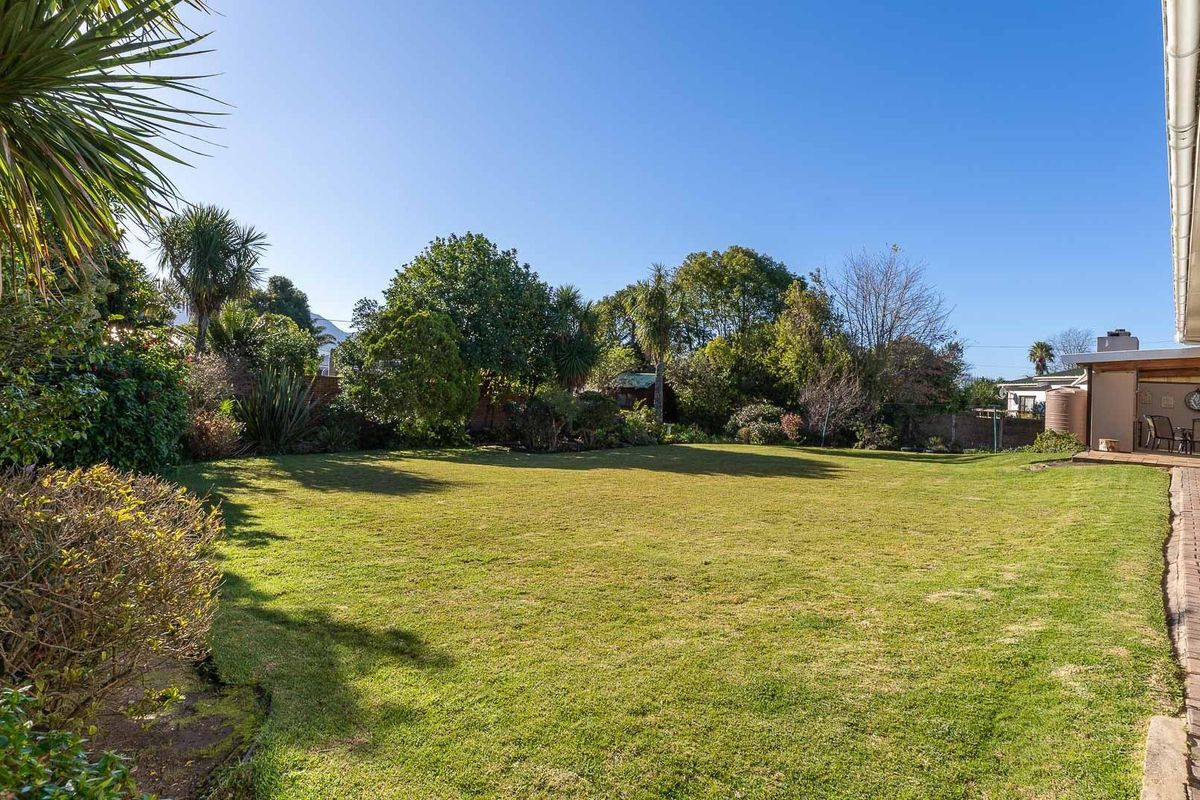 A spacious rear garden featuring a well-maintained lawn, mature landscaping, and a paved walkway leading to a covered patio area.