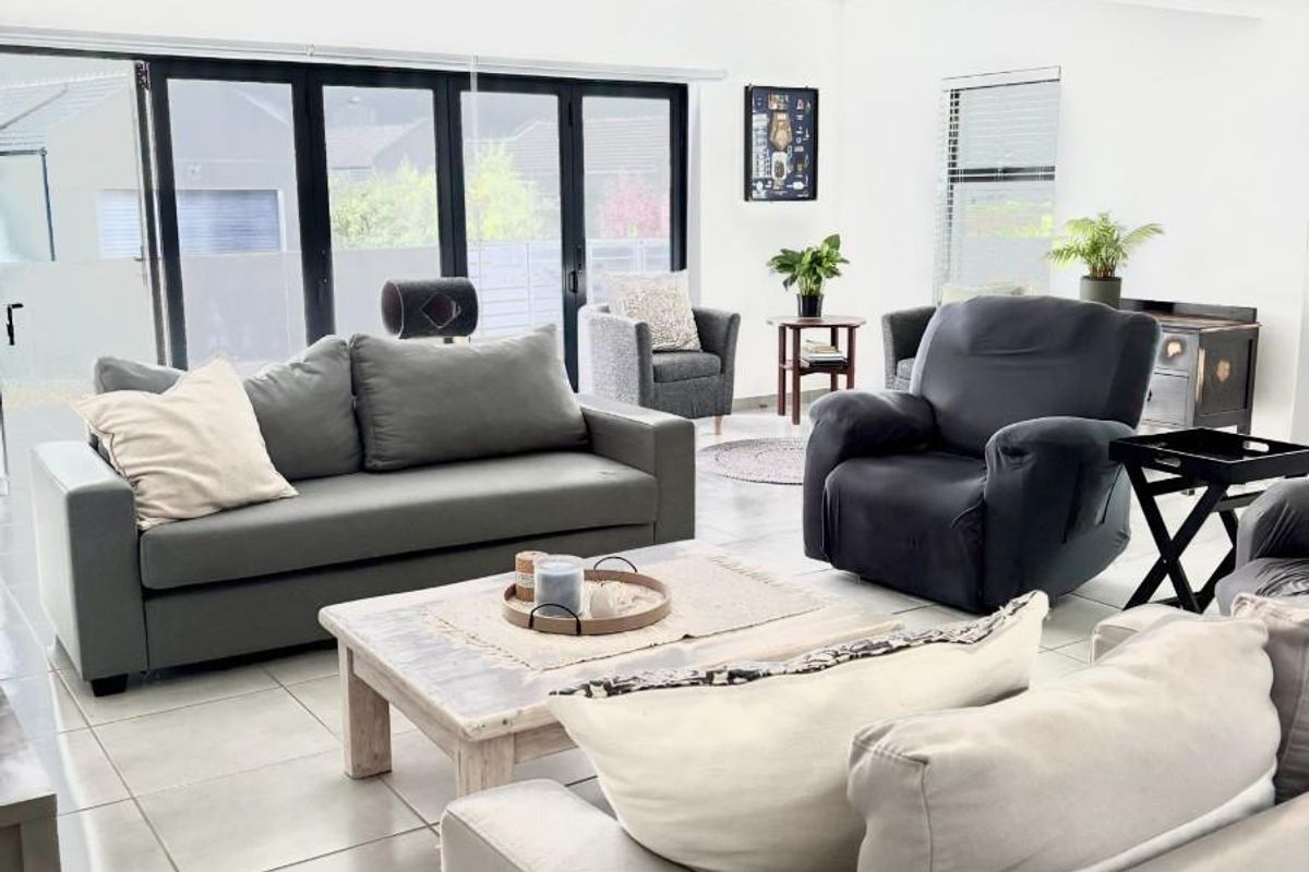 Spacious living room featuring light-colored tiled flooring, vaulted ceilings with exposed beams, and large black aluminium bifold doors providing ample natural light.
