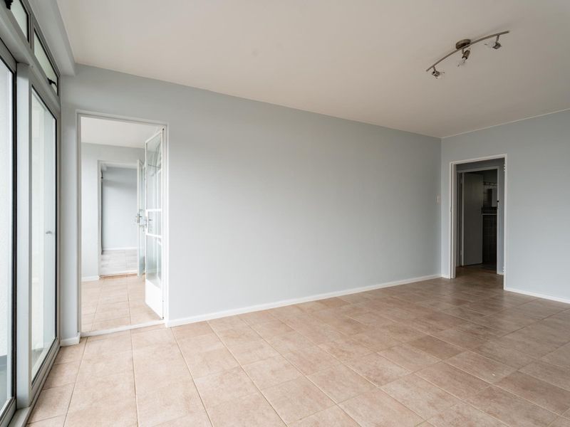 A bright, empty living area featuring neutral tiled flooring, track lighting, and access to an adjacent room through a glass-paneled door.