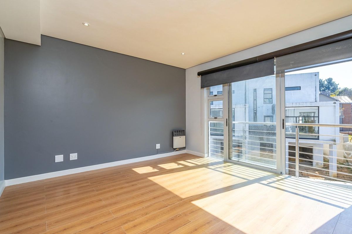 A bright, empty bedroom featuring laminate flooring, recessed lighting, and a sliding glass door leading to a private balcony with metal railings.