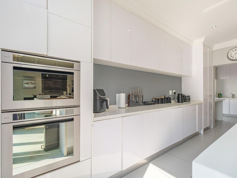 Modern kitchen featuring sleek white gloss cabinetry, integrated double ovens, stone countertops, and a seamless transition to the adjacent scullery area.
