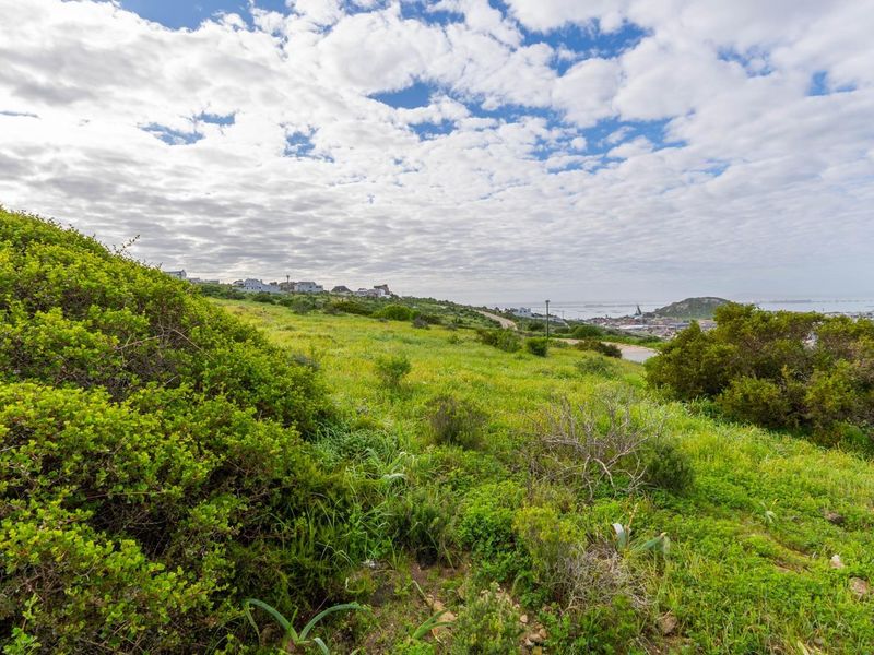 A scenic view overlooking a sloping grassy residential plot with distant coastal and neighborhood vistas under a cloudy sky.