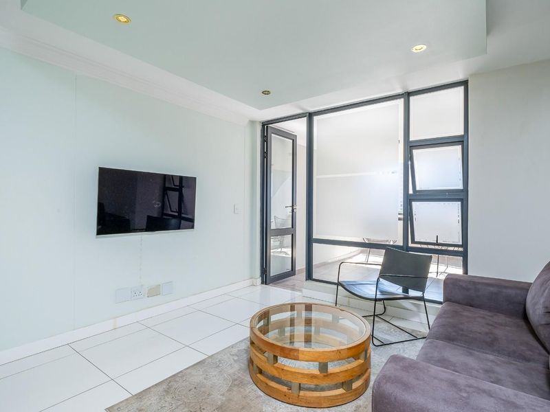 A bright and modern living room featuring tiled floors, recessed lighting, and direct access to an enclosed balcony through large aluminium-framed glass doors.