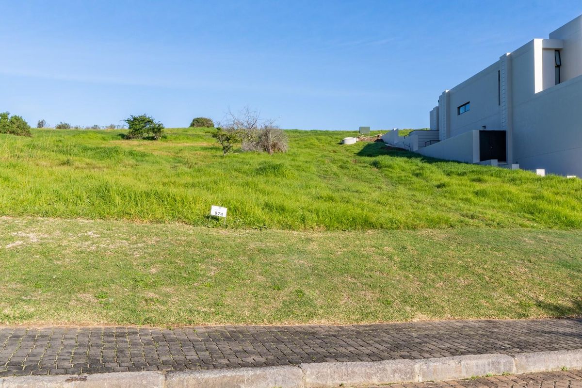 A vacant residential plot featuring a gentle upward slope, situated adjacent to an existing modern property with paved street frontage.