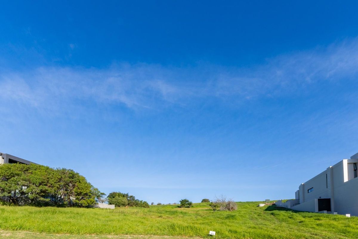 A view of a vacant residential plot featuring sloped grassy terrain, situated between existing modern residential properties under a clear blue sky.