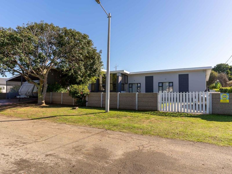 Front view of the property featuring a paved driveway, carport, and a secure boundary wall with a white picket gate.