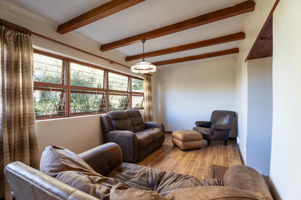A living room featuring exposed timber ceiling beams, laminate flooring, and large timber-framed windows with security bars, offering a rustic and cozy atmosphere.