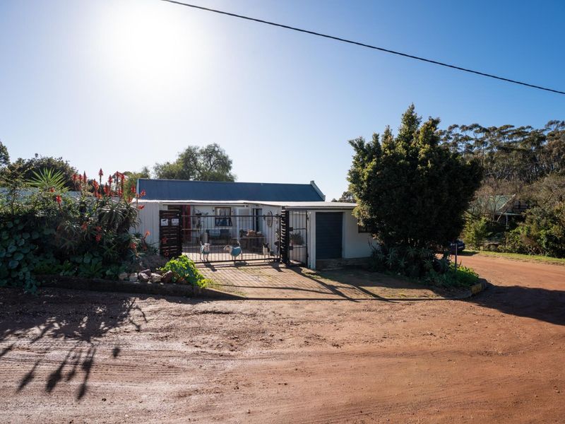 Front exterior view of a single-story residence featuring a corrugated metal roof, security gate, and driveway access from a dirt road.