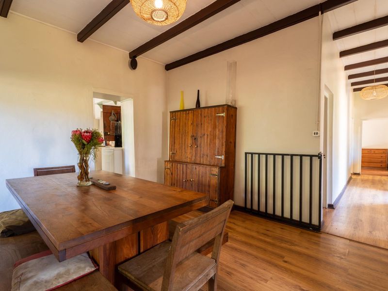 Dining area featuring exposed timber ceiling beams, laminate flooring, and a metal safety gate, with a view through to the kitchen and hallway.