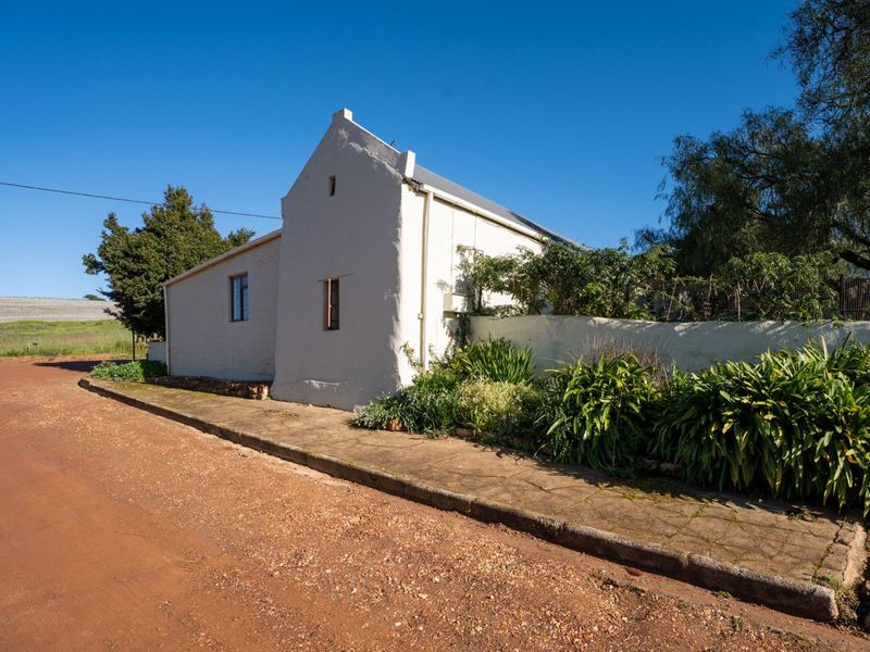 A view of the front facade of a traditional house with a gable roof, situated along a dirt road with a concrete sidewalk.