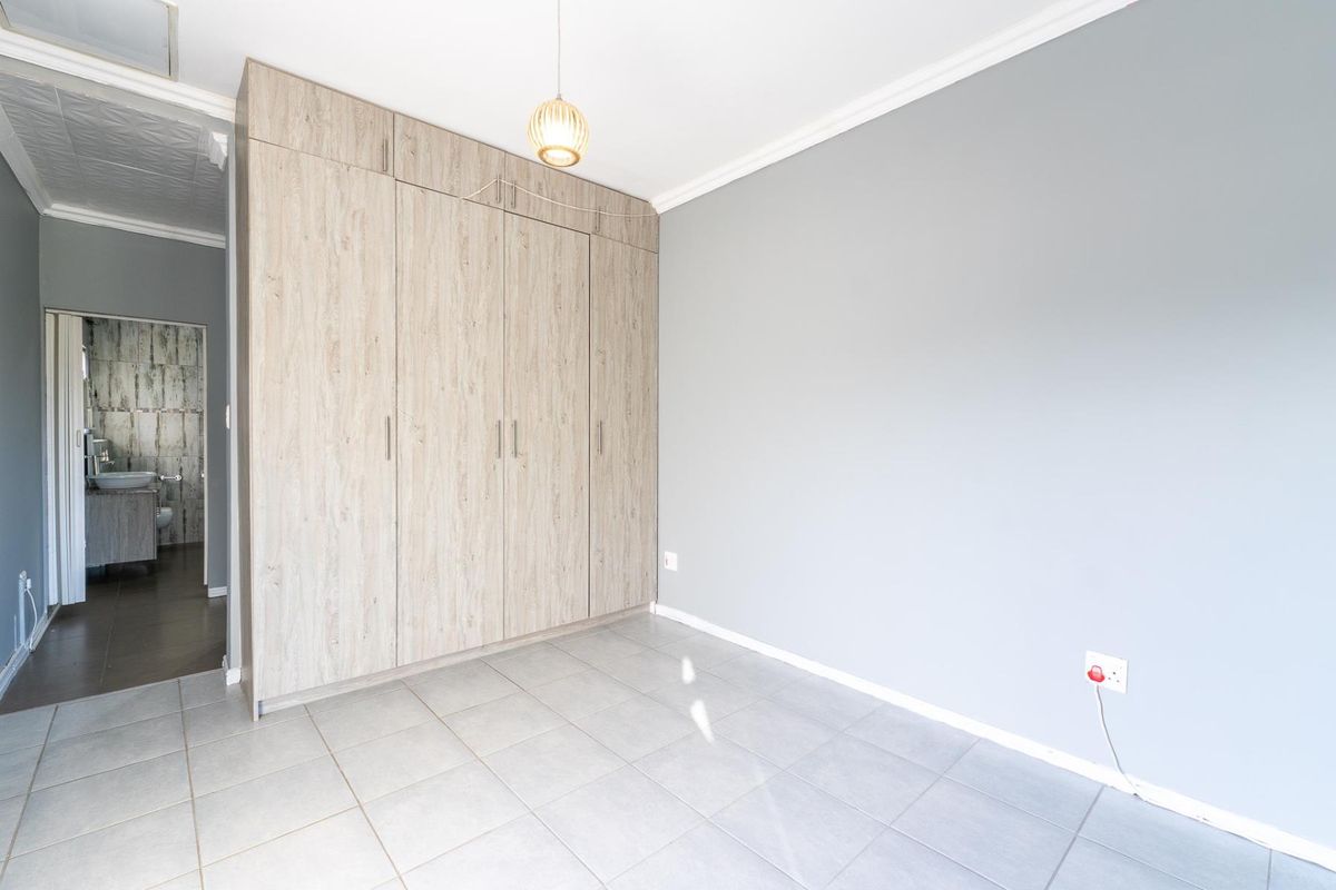 A bright, empty bedroom featuring neutral grey walls, light wood-grain built-in cupboards, and tiled flooring, with an adjacent view into a tiled bathroom.