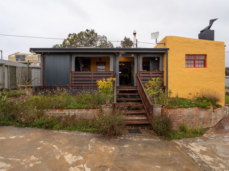 Front elevation of a house featuring a timber deck, staircase, and a mix of yellow rendered walls and dark corrugated metal cladding.