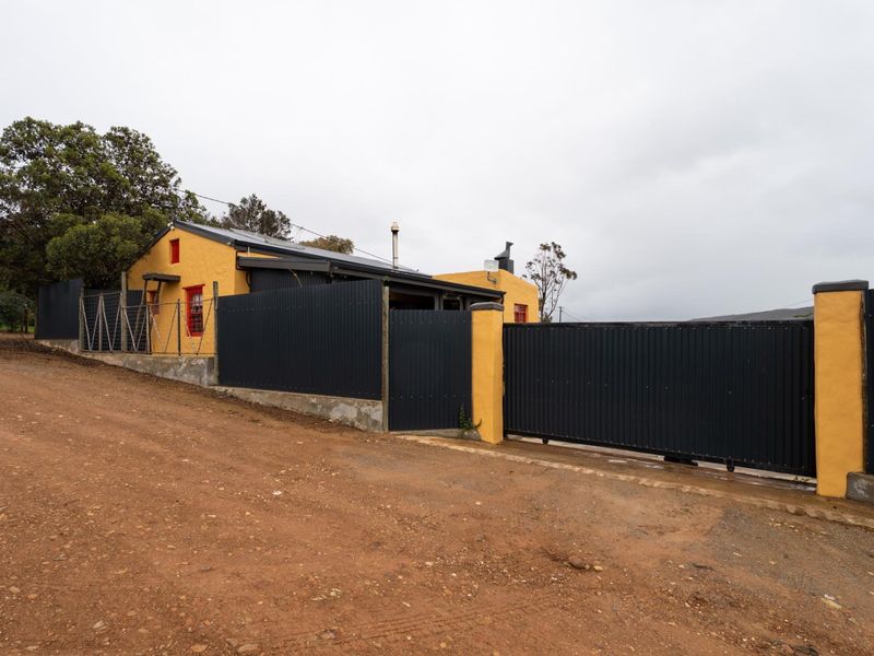Front exterior view of the property featuring a yellow facade, corrugated metal fencing, and a sliding gate entrance from an unpaved road.