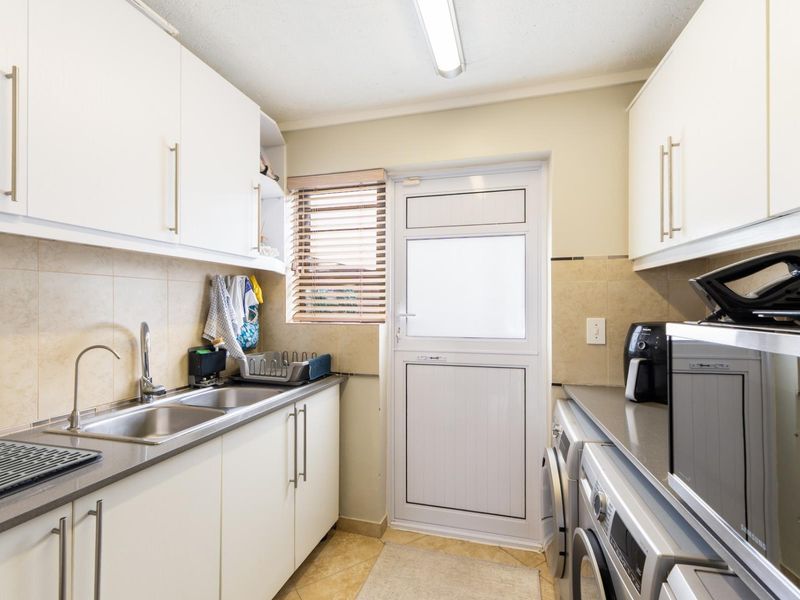 A functional scullery featuring white built-in cabinetry, a double stainless steel sink, tiled backsplash, and direct access to the exterior via a uPVC door.