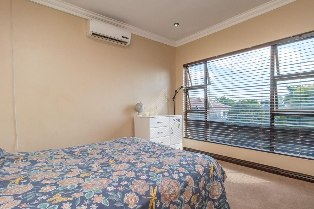 A well-lit bedroom featuring neutral walls, carpet flooring, a split-system air conditioner, and a large window with venetian blinds.