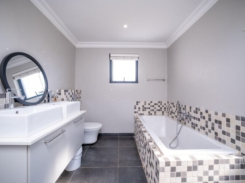 Modern bathroom featuring a double vanity, built-in bathtub with mosaic tile surround, dark floor tiles, and neutral wall finishes.