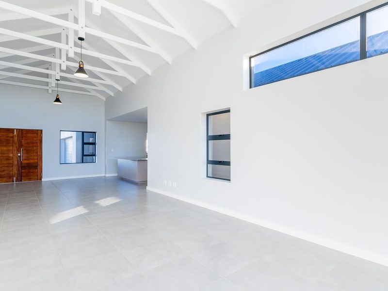 Spacious open-plan living area featuring high vaulted ceilings with exposed trusses, modern tiled flooring, and abundant natural light through large windows.