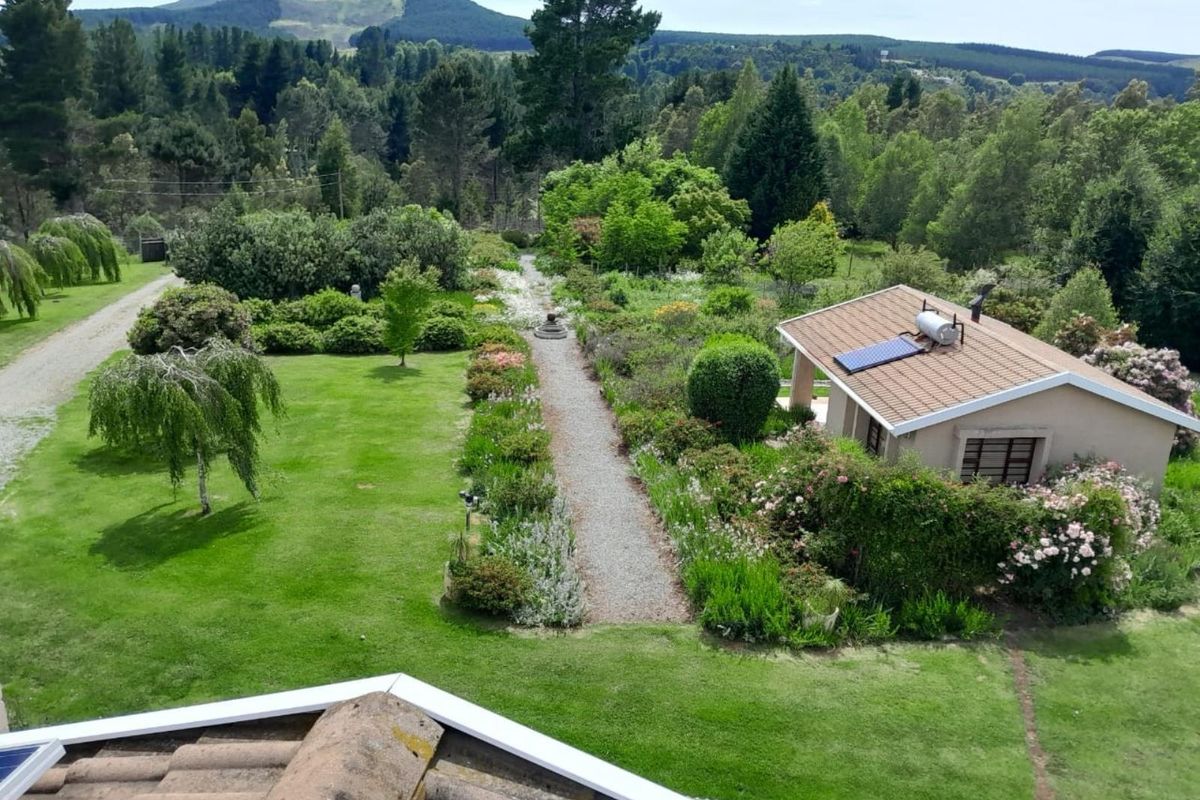 An elevated aerial view of a lush, landscaped garden featuring a gravel pathway, mature trees, and a secondary building with a solar water heater.