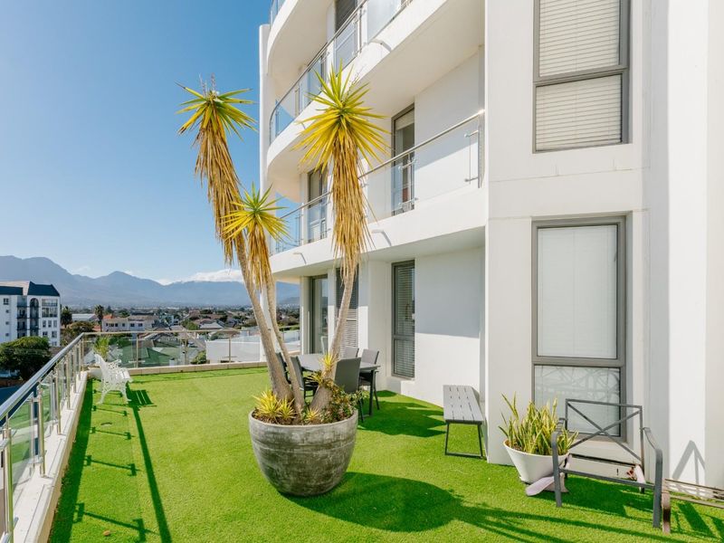 A spacious apartment balcony featuring artificial turf flooring, glass balustrades, and expansive views of the surrounding neighborhood and mountains.
