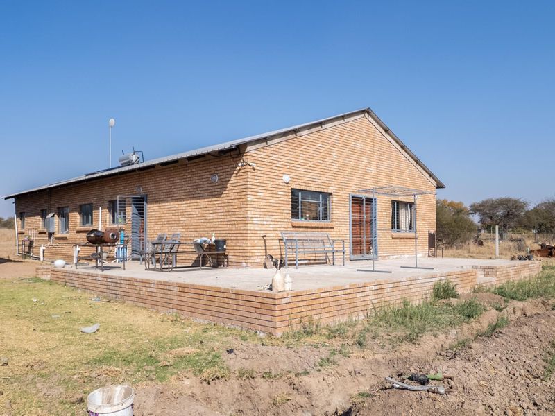 Rear view of a face brick farmhouse featuring a spacious concrete patio, solar water heating system, and metal roofing in a rural setting.
