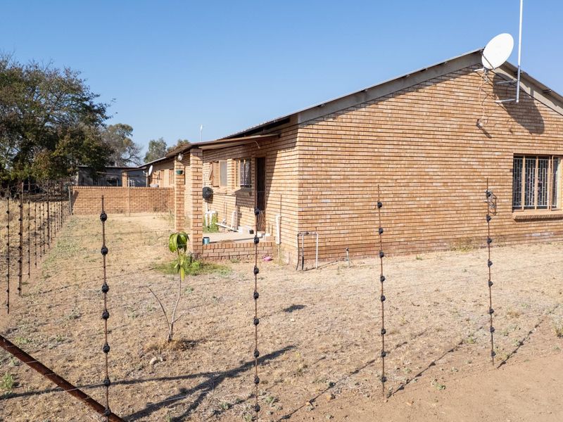 Rear view of a face brick farm dwelling featuring security bars, a satellite dish, and an enclosed yard with electric fencing.