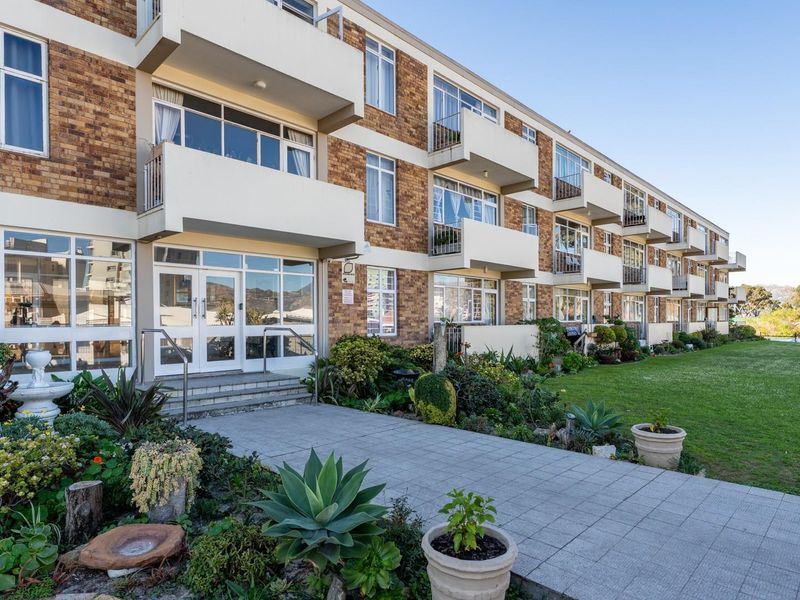 A well-maintained apartment building exterior featuring a brick facade, private balconies, a paved entrance walkway, and established garden landscaping.