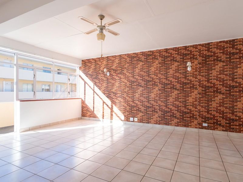 Spacious, sunlit living area featuring ceramic floor tiles, a ceiling fan, and a prominent exposed brick feature wall.