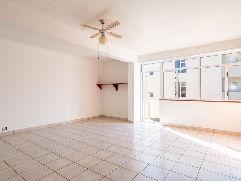 A bright, empty living room featuring tiled floors, a ceiling fan, and a brick-effect feature wall with large windows providing natural light.