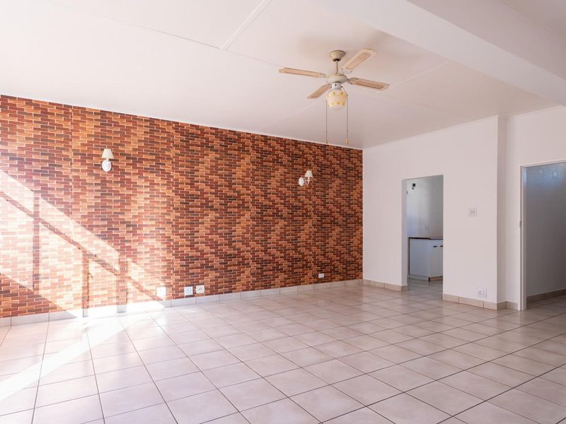 Spacious living area featuring a brick-style accent wall, neutral ceramic floor tiles, and a ceiling fan, offering a bright and versatile open-plan layout.