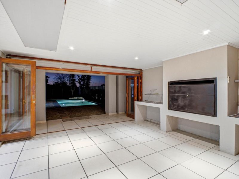 Spacious covered patio featuring a built-in braai, tiled flooring, and large wooden folding doors opening out to the swimming pool area.