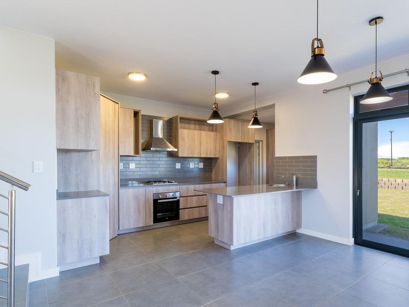 Modern open-plan kitchen featuring light wood-grain cabinetry, stone countertops, integrated stainless steel appliances, and direct access to the rear garden through sliding doors.