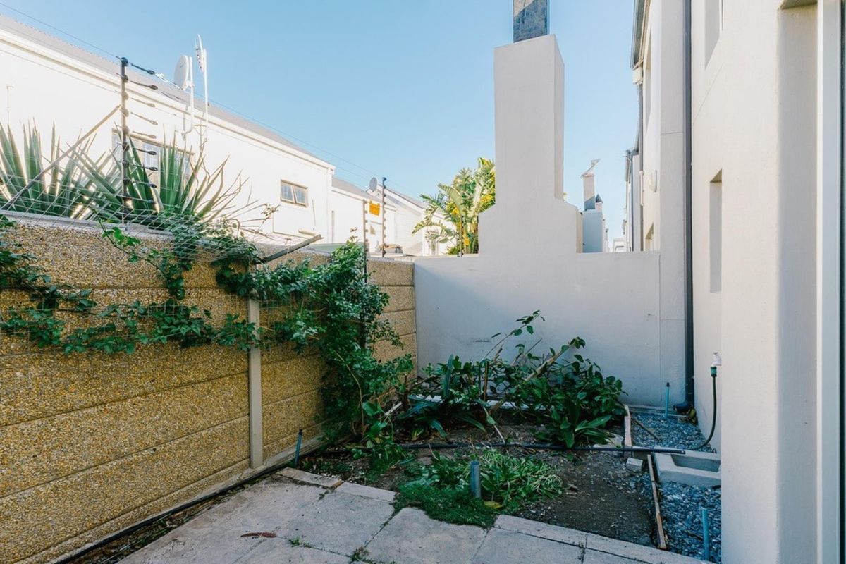 A compact rear courtyard featuring a paved patio, boundary wall with electric fencing, and an external chimney, requiring landscaping and maintenance.