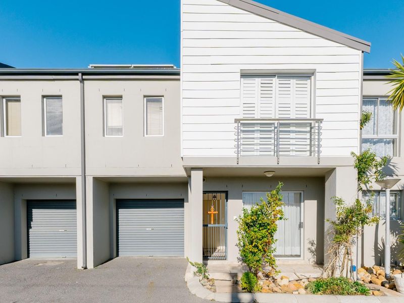 Front elevation of a modern townhouse featuring a paved driveway, roller garage doors, and a private balcony with metal railings.