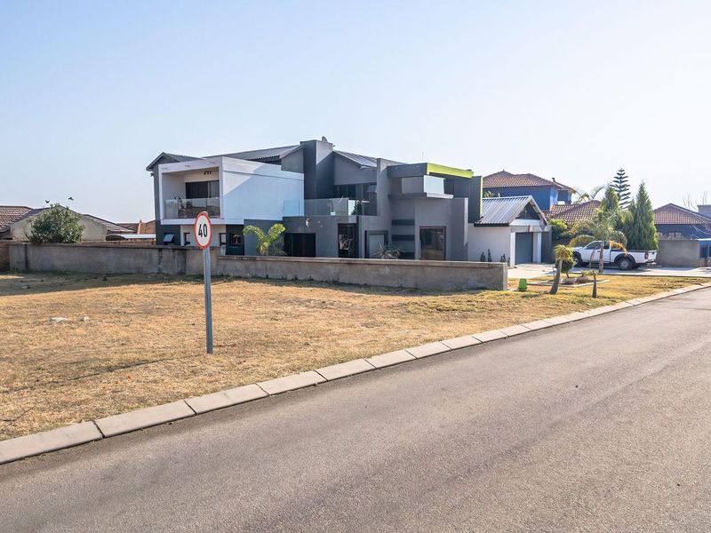 Street view of a vacant residential plot adjacent to a modern multi-story home with a concrete boundary wall and paved road access.