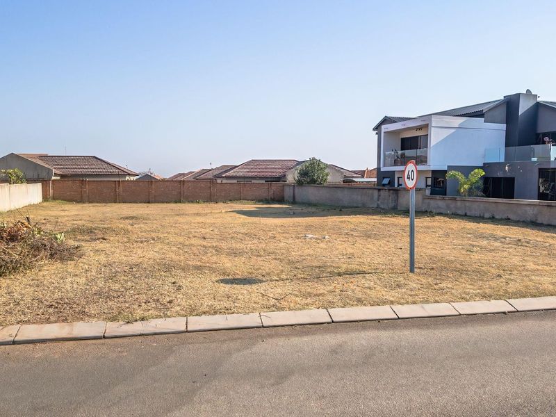 A vacant residential plot of land featuring a perimeter brick wall, situated along a paved road in a developed neighborhood.