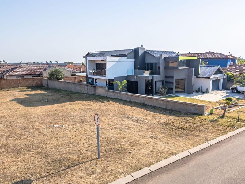An aerial view of a vacant residential plot situated next to a modern multi-story home, featuring a perimeter wall and street access.