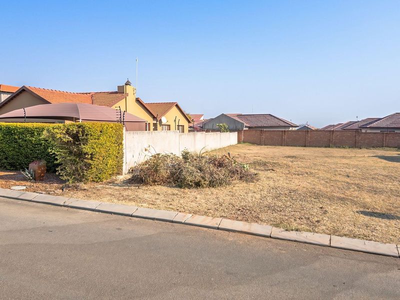 A vacant residential plot featuring a concrete boundary wall with electric fencing, situated along a paved street in a developed neighborhood.