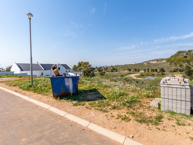A view of a vacant residential plot featuring a paved road, street lighting, and surrounding natural landscape.